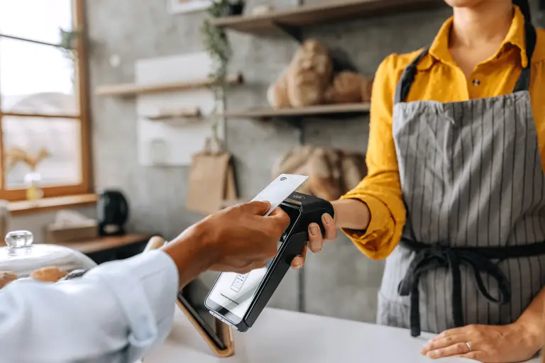 Un client règle son achat en approchant sa carte bancaire d’un terminal de paiement sans contact, tenu par une personne portant un tablier dans une boulangerie.